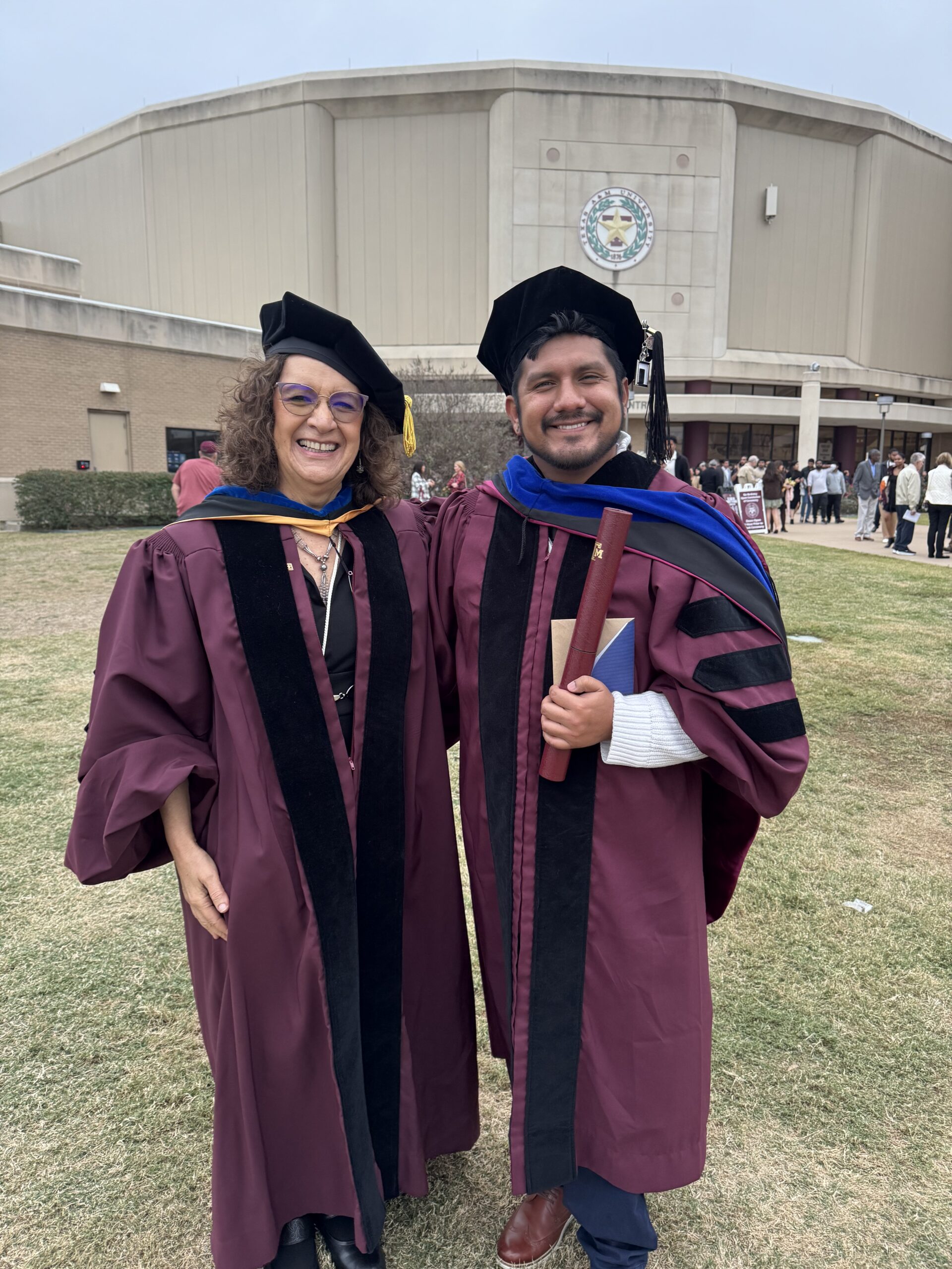 Dr. Patricia Pietrantonio and Jonathan Hernandez, Ph.D., both wearing doctoral hoods and standing in front of reed arena following the graduation for Jonathan.